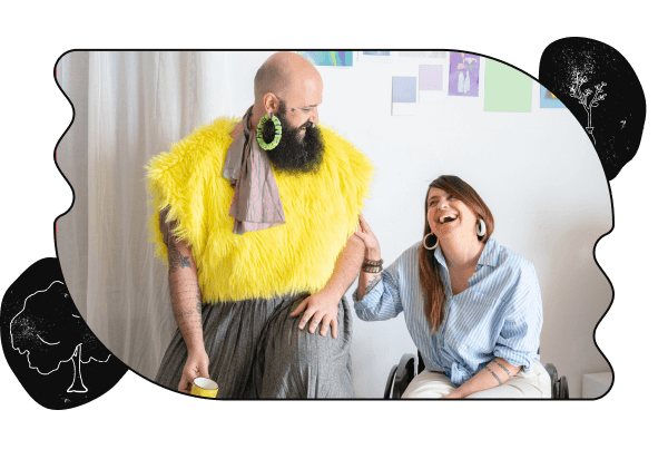 A picture of two people talking and laughing. The person on the left has a beard and is wearing a skirt and fuzzy yellow top. The person on the right is touching their arm while laughing and is sitting in a wheelchair. The image is cropped in a bubbly shape with a black border and tree sketches on the top-left and bottom-right corners.