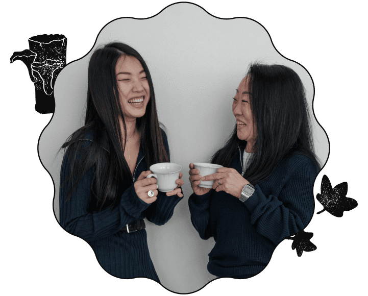 An image of two east-asian women laughing and holding coffee cups. The image is cropped to a wavy-shaped frame with a black border, with a stamp of two mushrooms growing out of a log on the top-left and a stamp of two leaves on the bottom-right.